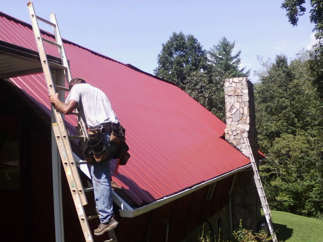 Roofer on a ladder beside a red metal roof and stone chimney — Soffit & Fascia Repair by Roofing Brothers in Matthews